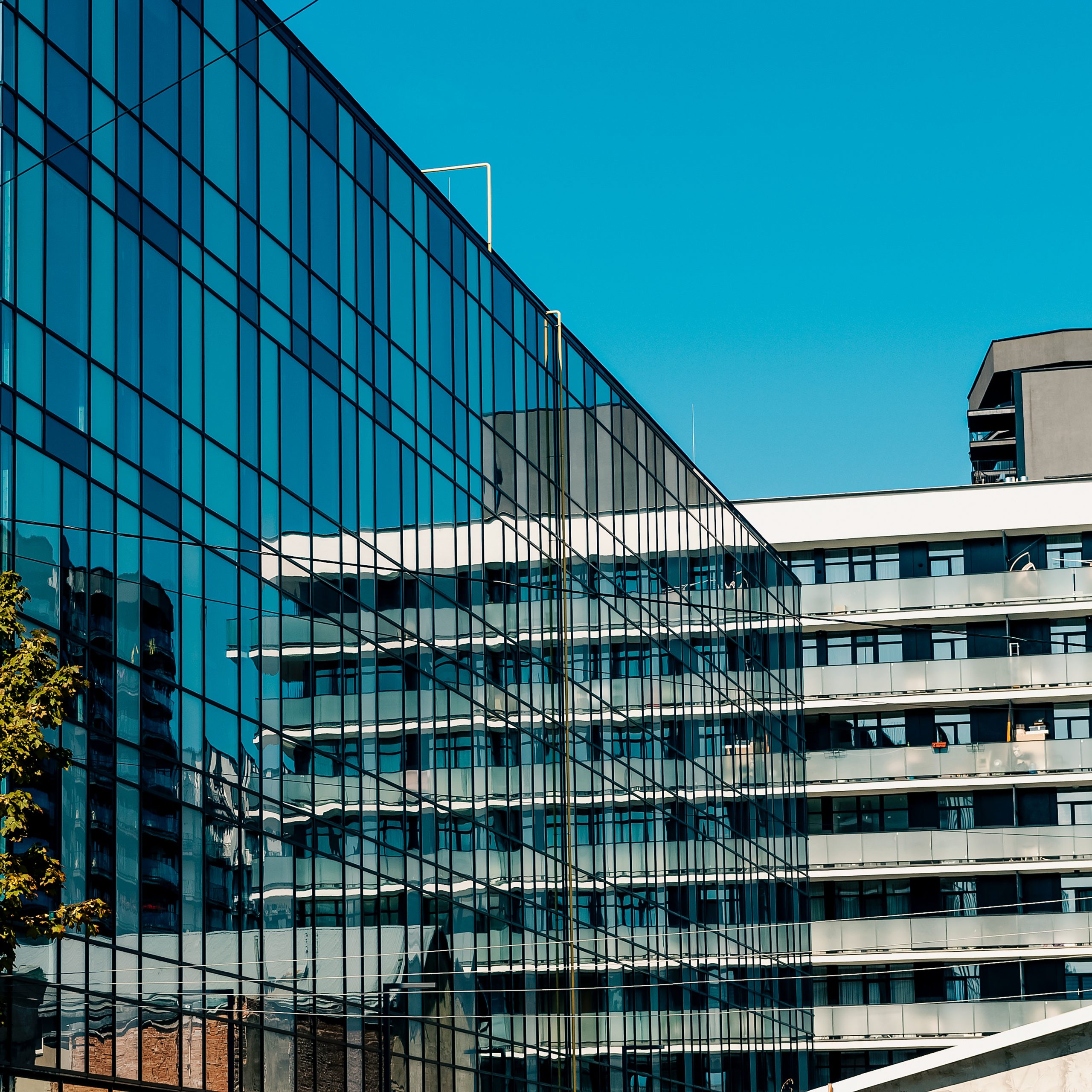 Fragment nowoczesnego kompleksu budynków, na pierwszym planie duża fasada biurowca w całości pokryta szkłem, odbijająca błękitne niebo i architekturę sąsiedniego budynku. Drugi budynek w tle ma szarą elewację z rzędami balkonów ze szklanymi i metalowymi balustradami.