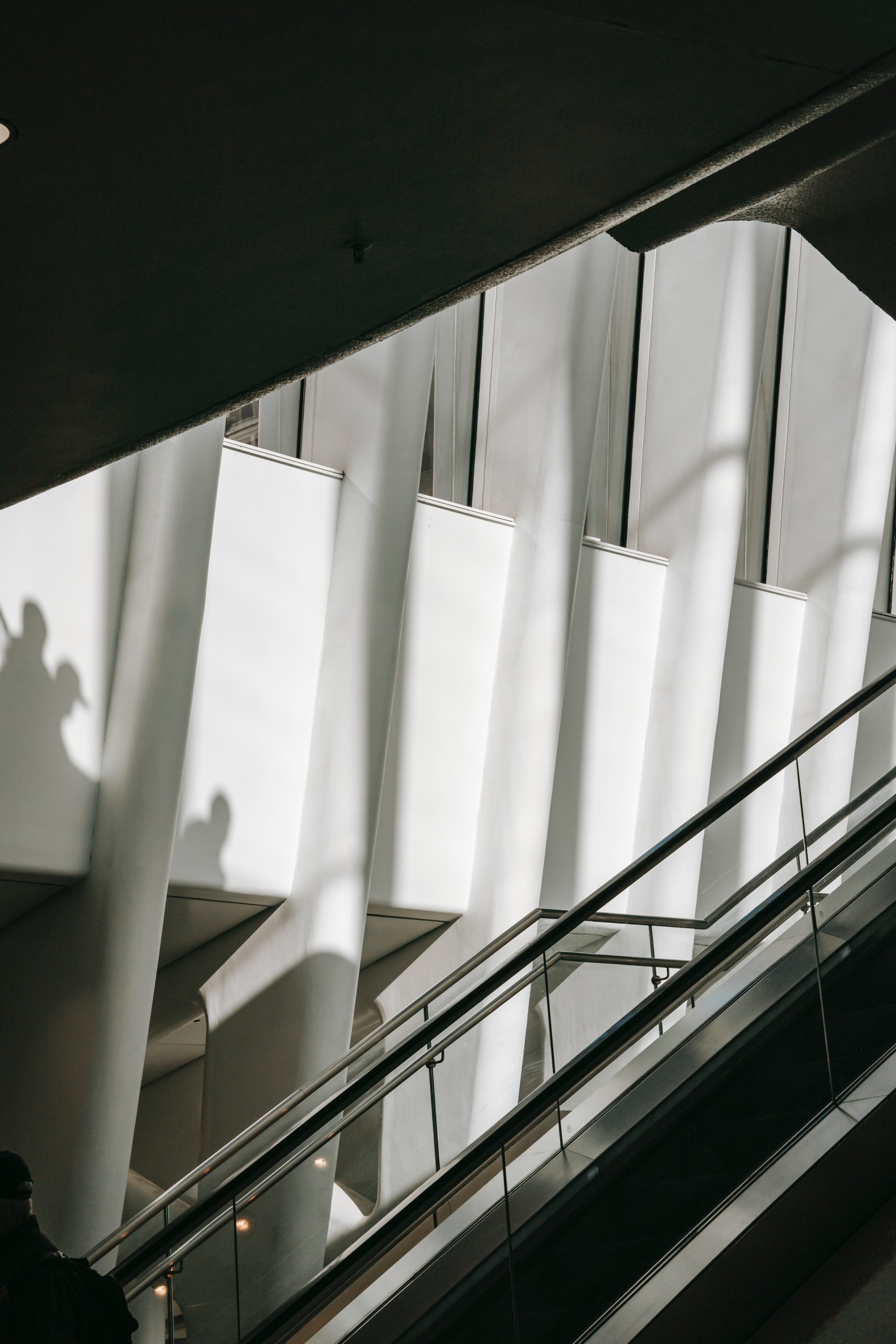 Fragment nowoczesnego wnętrza, najprawdopodobniej na lotnisku lub w centrum handlowym, ukazujący biegnące po przekątnej schody ruchome ze szklaną i metalową balustradą. Nad schodami widoczna jest zakrzywiona ściana z pionowymi, białymi panelami, rzucająca głębokie cienie. Na ścianie widoczne są cienie sylwetek ludzi stojących wyżej.