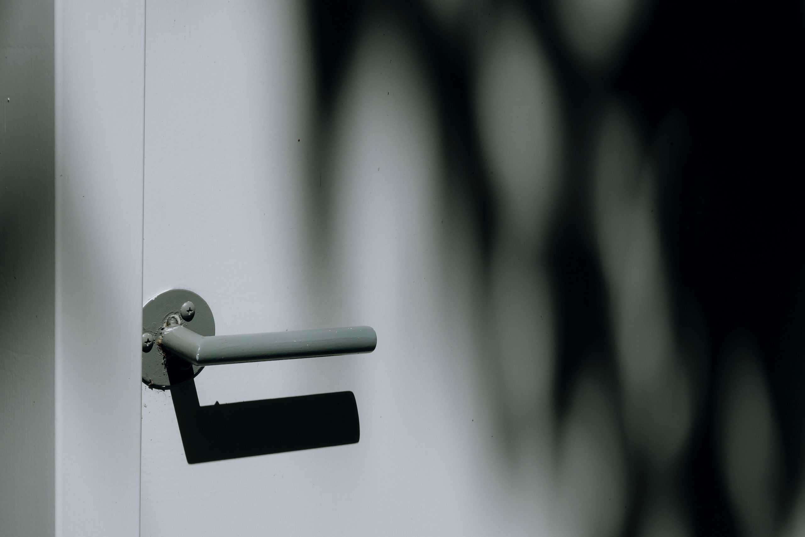 Zbliżenie na minimalistyczną, metalową klamkę pomalowaną na szaro lub jasnozielono, zamontowaną na białych lub jasnoszarych drzwiach. Na drzwiach widoczny jest wyraźny cień rzucany przez klamkę oraz rozmyte, duże cienie liści lub gałęzi po prawej stronie.