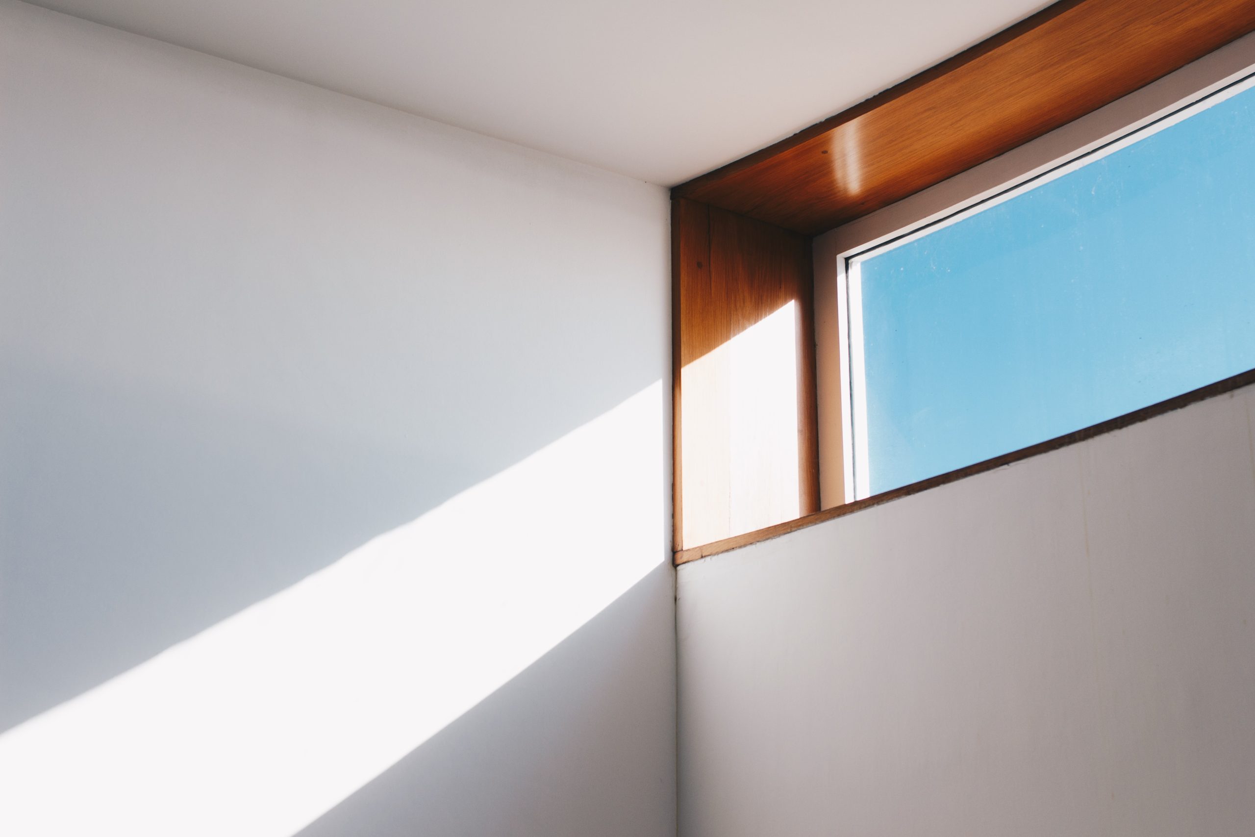 Minimalistyczne ujęcie wnętrza z białymi ścianami, na które pada ostry, diagonalny cień. W prawym górnym rogu znajduje się poziome okno z drewnianą, łukowatą ramą, przez które widać błękitne niebo.