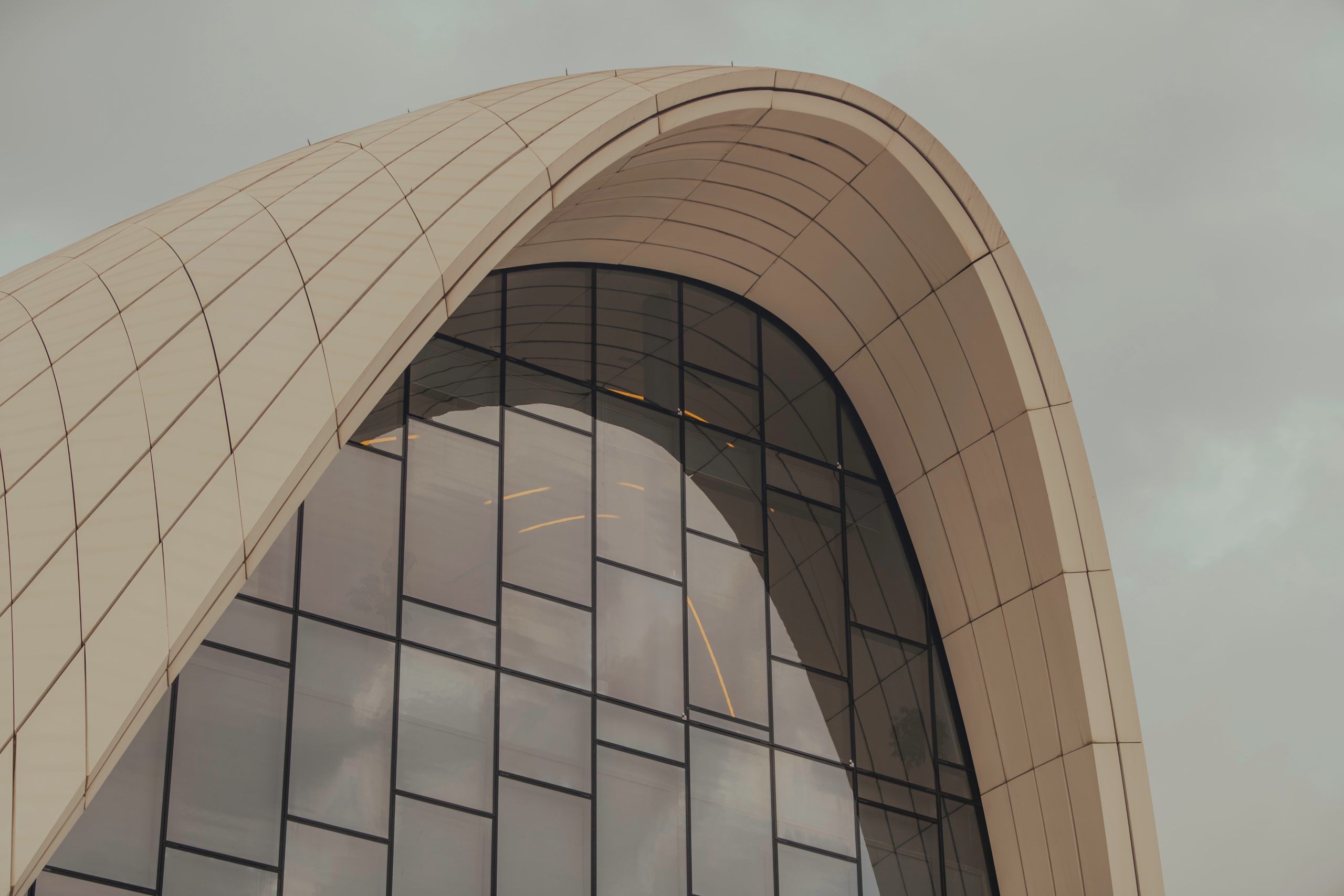 Zbliżenie na fragment futurystycznej architektury, charakteryzującej się płynnym, zakrzywionym dachem/fasadą wykończoną beżowymi panelami. Pod spodem znajduje się duża, łukowata szklana ściana, podzielona na geometryczne segmenty czarnymi ramami. Architektura przypomina budynek Heydar Aliyev Center w Baku. Tło stanowi jednolite, szare niebo.