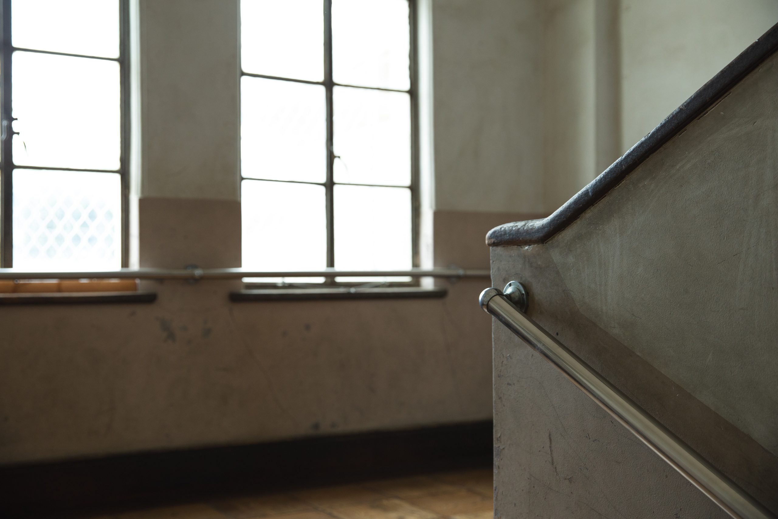 Fragment starego, ascetycznego wnętrza, prawdopodobnie klatki schodowej lub korytarza. Na pierwszym planie widoczny jest masywny betonowy lub tynkowany murek schodów z metalową, minimalistyczną balustradą. W tle znajdują się dwa prostokątne okna z podziałami szprosowymi, wpuszczające jasne, rozproszone światło. Ściany są brudne i zaniedbane.