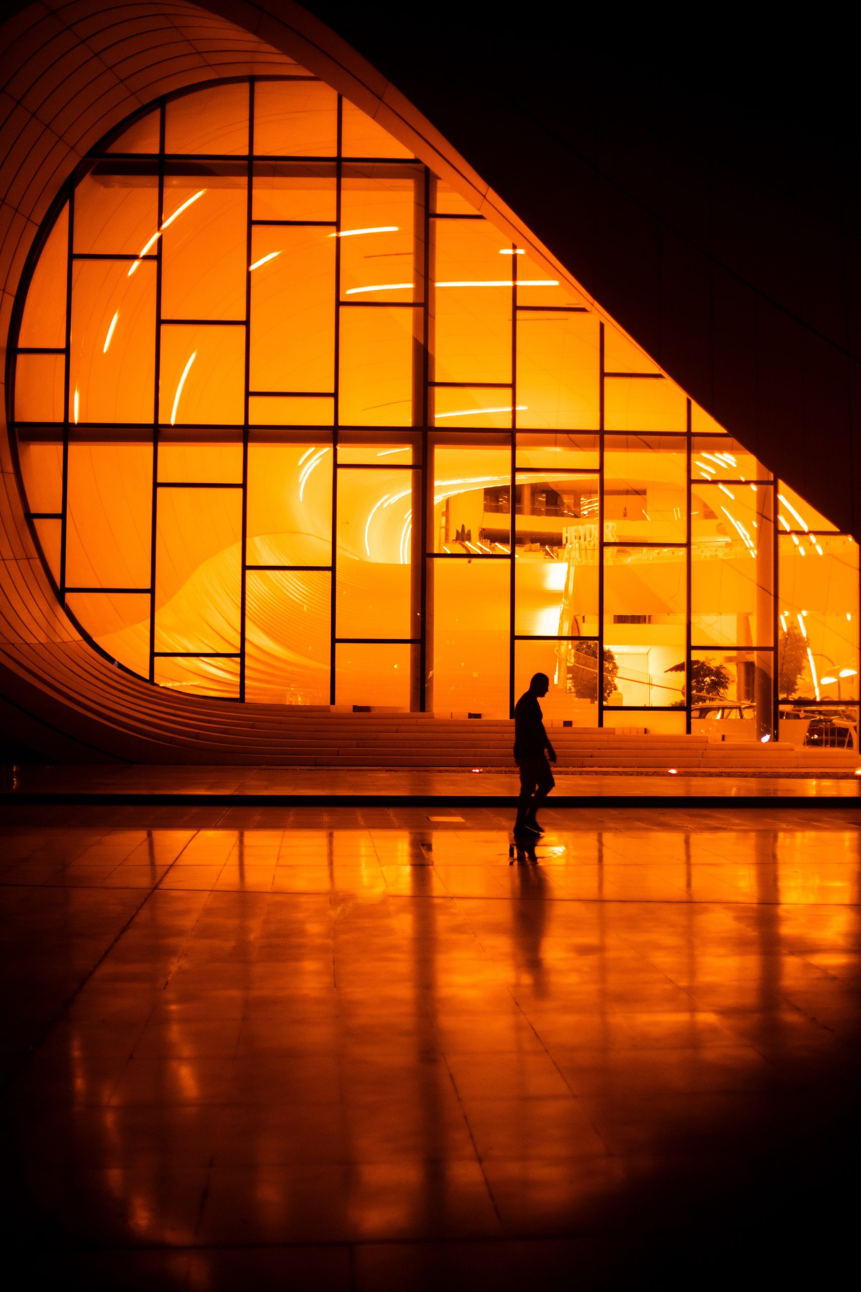 Nocne ujęcie futurystycznej fasady budynku, prawdopodobnie Heydar Aliyev Center w Baku, charakteryzującej się obłymi kształtami i dużą, siatkowaną szklaną ścianą. Wnętrze jest intensywnie oświetlone pomarańczowym lub bursztynowym światłem. Na gładkiej, błyszczącej podłodze na pierwszym planie widoczna jest sylwetka pojedynczej osoby, której cień i odbicie są widoczne na posadzce.